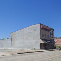 A flat-fronted, grey commercial building stands beside a smaller, multi-story structure under a clear, bright blue sky.