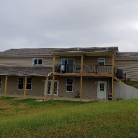 Two-story house with a wooden deck and covered porch, featuring tan siding and gray roof shingles over a grassy yard.