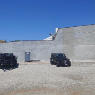 A black Jeep and a trailer are parked on a gravel lot in front of a long, gray concrete wall under a clear blue sky.