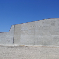 A large, grey concrete wall stands against a clear, bright blue sky above a light-colored gravel ground.