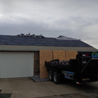 A residential garage with a roof under repair, featuring underlayment and a dump trailer parked in the driveway.