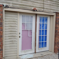 A set of exterior French doors with white frames and mismatched panel inserts, next to a panel of weathered beige siding.