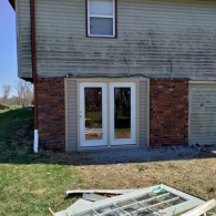 New white double doors installed in a brick and siding wall of a house, with an old discarded door lying in the grass.
