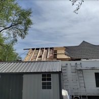 Construction workers replacing a residential roof, with exposed rafters and plywood visible on an unfinished section.