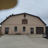 A large tan barn-style building with two doors, two windows, and a garage door, featuring a dark brown roof and trim.