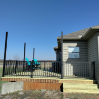 A wooden deck featuring black railing and tall posts, set against a house with tan siding under a clear blue sky.