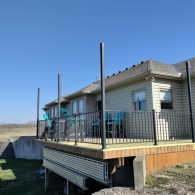 A tan house with a wooden deck featuring black railings and tall, unfinished support posts against a clear blue sky.