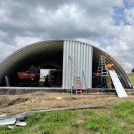 Construction of a corrugated metal Quonset-style shed, with a worker on a ladder installing a metal panel.