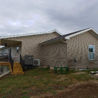 A tan, single-story house with a wooden staircase leading to a covered porch and a central air conditioning unit outside.