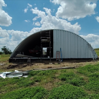 An unfinished Quonset hut building with corrugated metal siding, scaffolding, and a ladder under a cloudy blue sky.