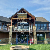 A two-story house with a rear deck and stone pillars, featuring metal scaffolding and a ladder set up for maintenance work.