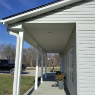 A view along a concrete front porch of a light gray house with white columns, soffit, and siding under a clear blue sky.