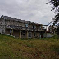 A two-story tan house with a covered wooden porch, set on a grassy, sloped hill under a cloudy sky.