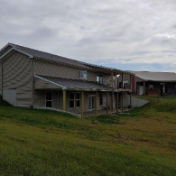 A beige two-story house with a wooden porch and a sloping grassy yard under a cloudy sky.