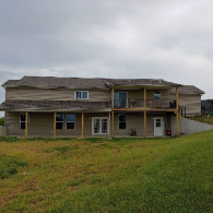 A two-story tan house with a large wooden deck and covered porch sits in a green, grassy yard under a cloudy sky.