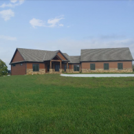 A single-story ranch house with a brown exterior, stone wainscoting, and a grey shingled roof, set on a green lawn.