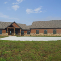 A single-story building with tan stone siding, wood trim, and a grey shingled roof, set behind a circular concrete driveway.