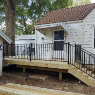 A new wooden deck with black metal railings built in front of a white house with a brown shingled roof.