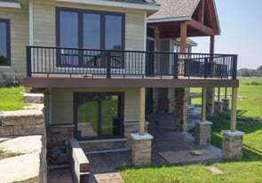 A tan house with a black-railed deck over a stone-paved patio with a basement window and a retaining wall.