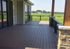 A wooden deck with black railings leads to sliding glass doors, overlooking a grassy field under a bright sky.