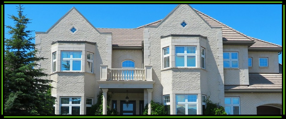 A light-colored, multi-story suburban home with a balcony and a tall evergreen tree to the left under a bright blue sky.