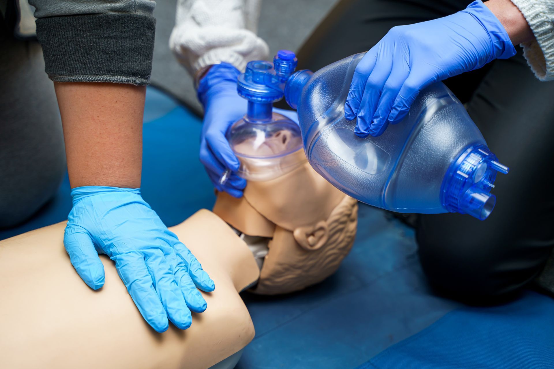 Two people in blue medical gloves perform CPR on a training mannequin using a bag valve mask. Two people in blue medical gloves perform CPR on a training mannequin using a bag valve mask.