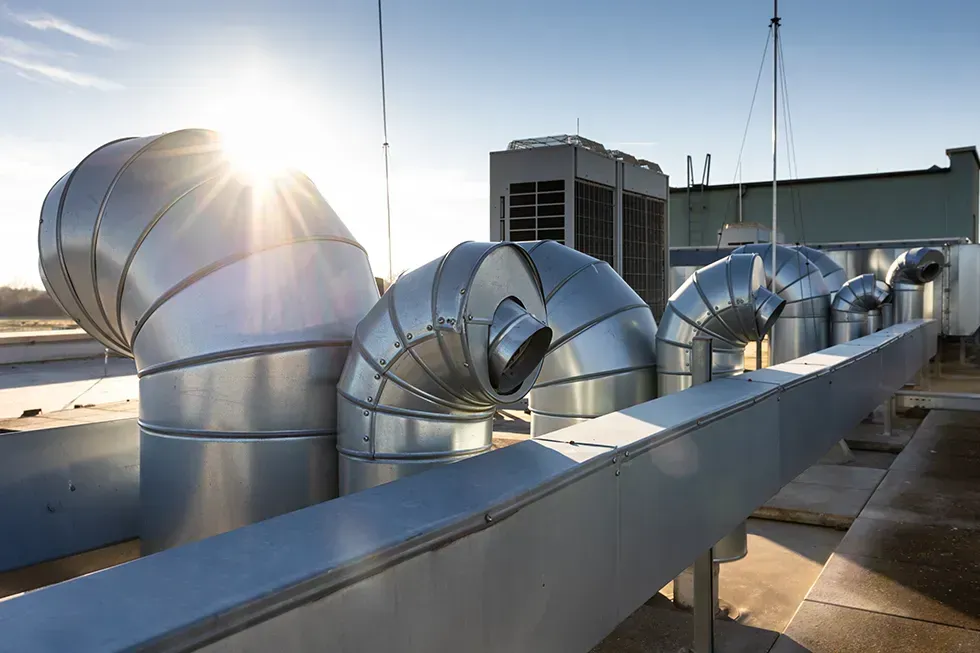 Rooftop HVAC system with silver ductwork, vents, and a cooling unit against a bright sun.