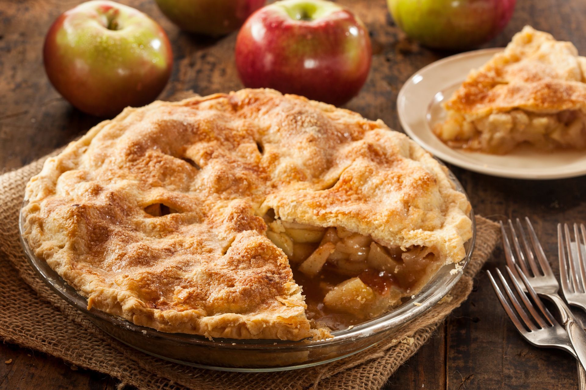Apple pie in a glass pie dish with a missing slice, several apples, and forks on a wooden table.