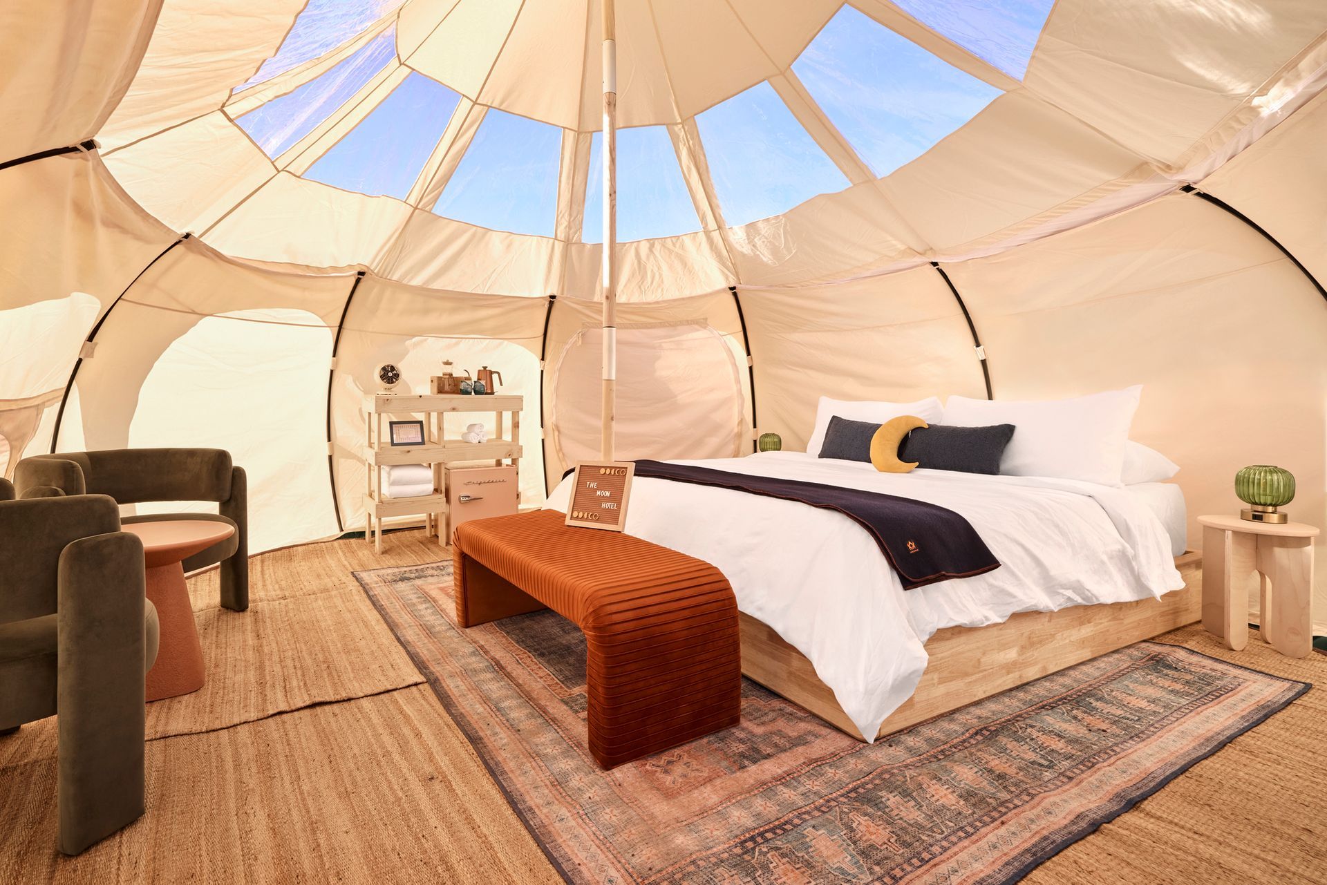 Inside a large glamping tent, a bed with a brown bench, sitting area, and skylights.