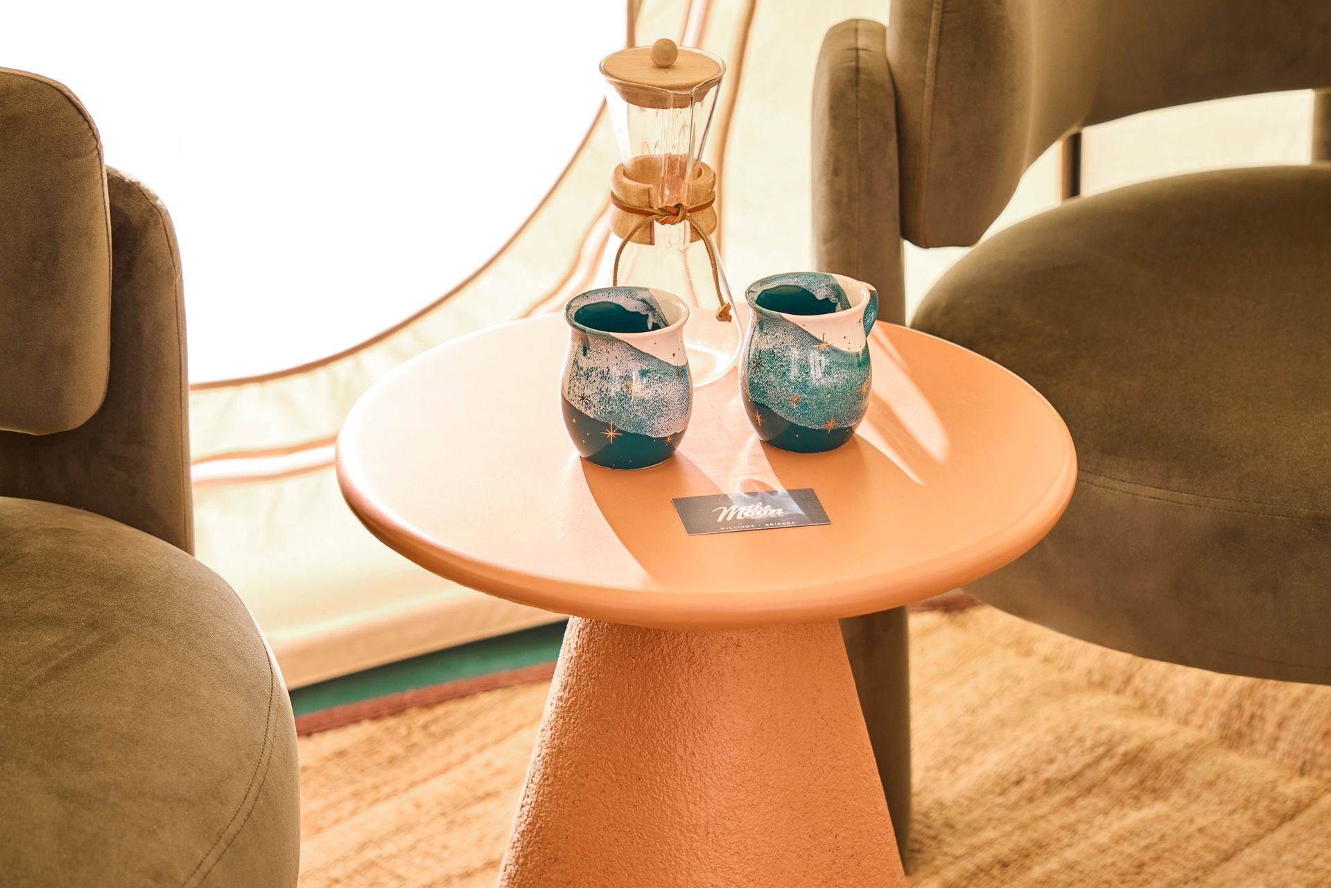 Orange table with two blue cups and a coffee maker, flanked by two gray chairs.