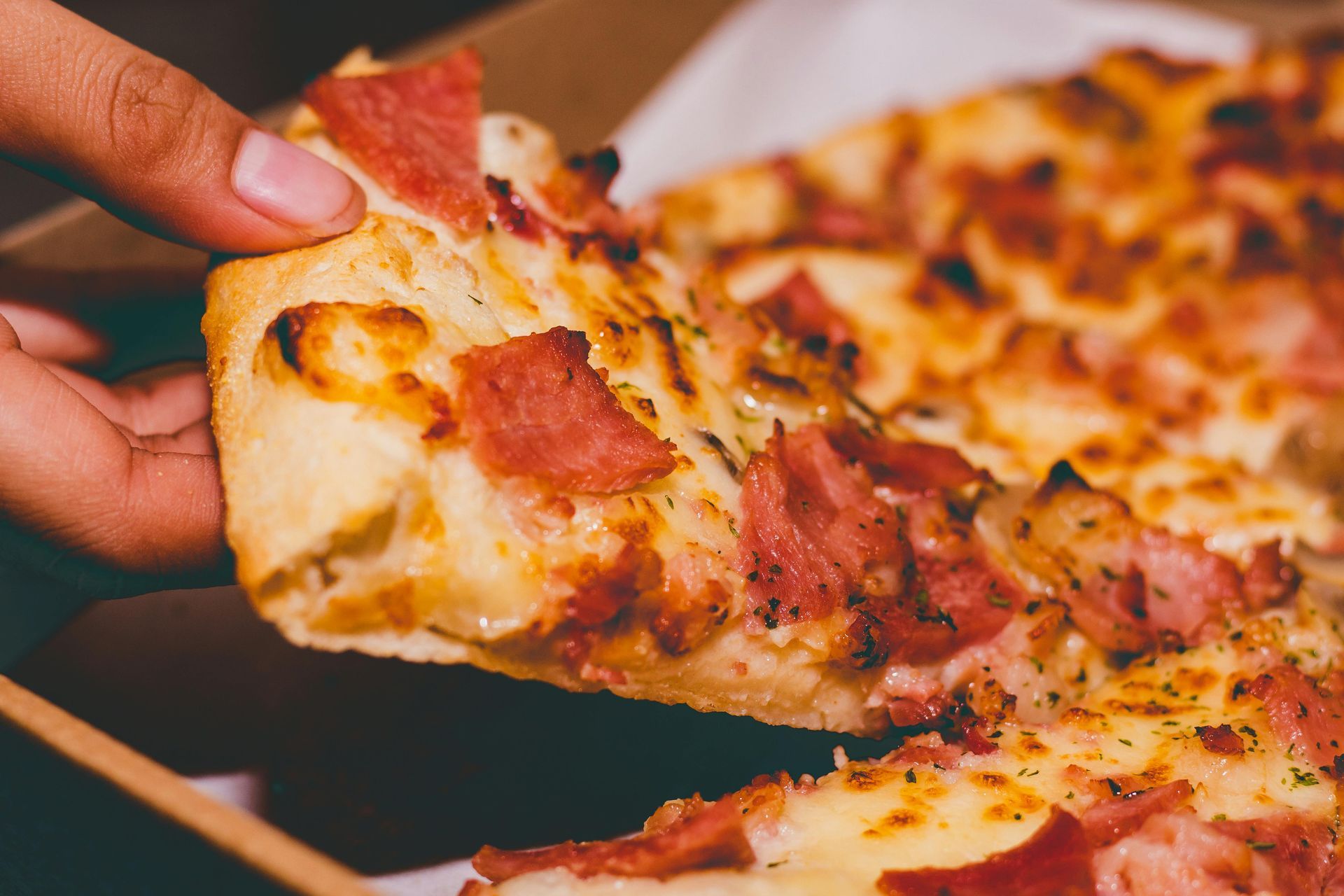 Hand holding a slice of pizza with ham and cheese. Pizza sits in a cardboard box.