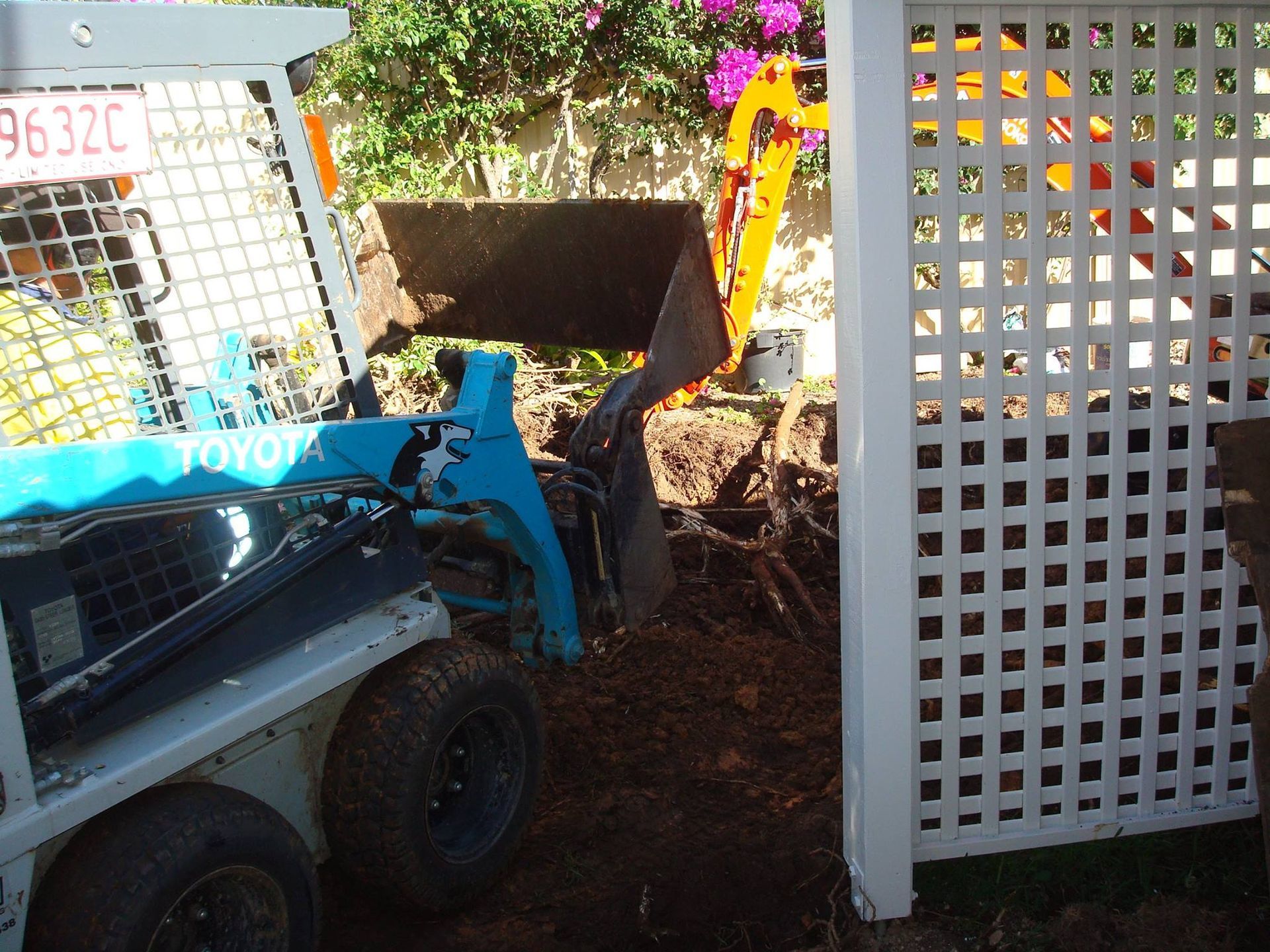 Blue Toyota skid steer loader digging near a white lattice fence, garden setting.