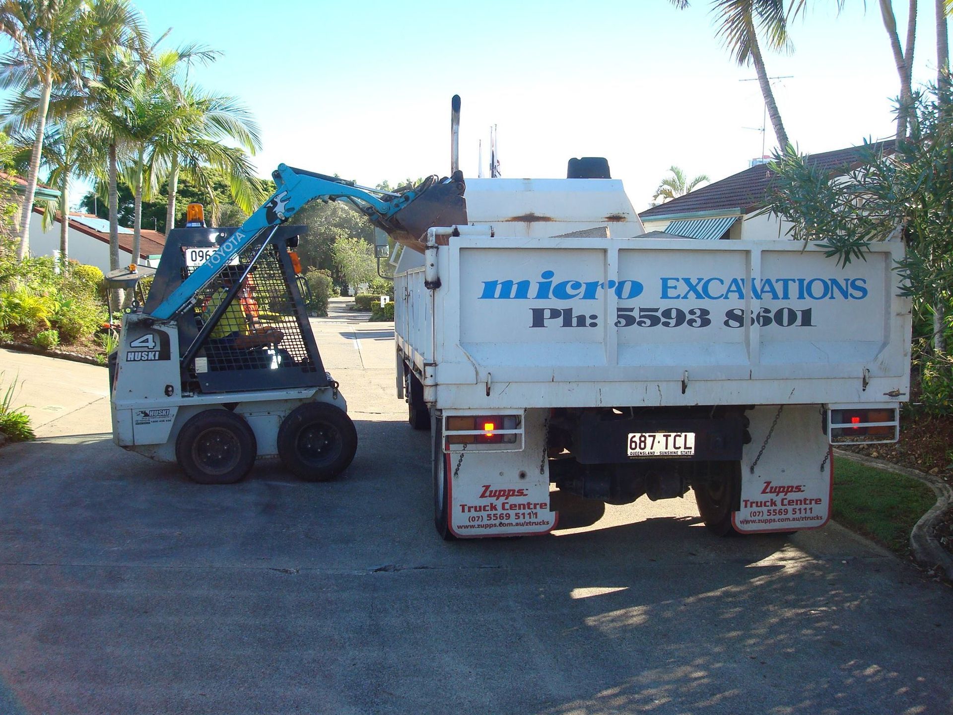 Micro excavator loading a white dump truck with soil. Outdoor setting with palm trees.