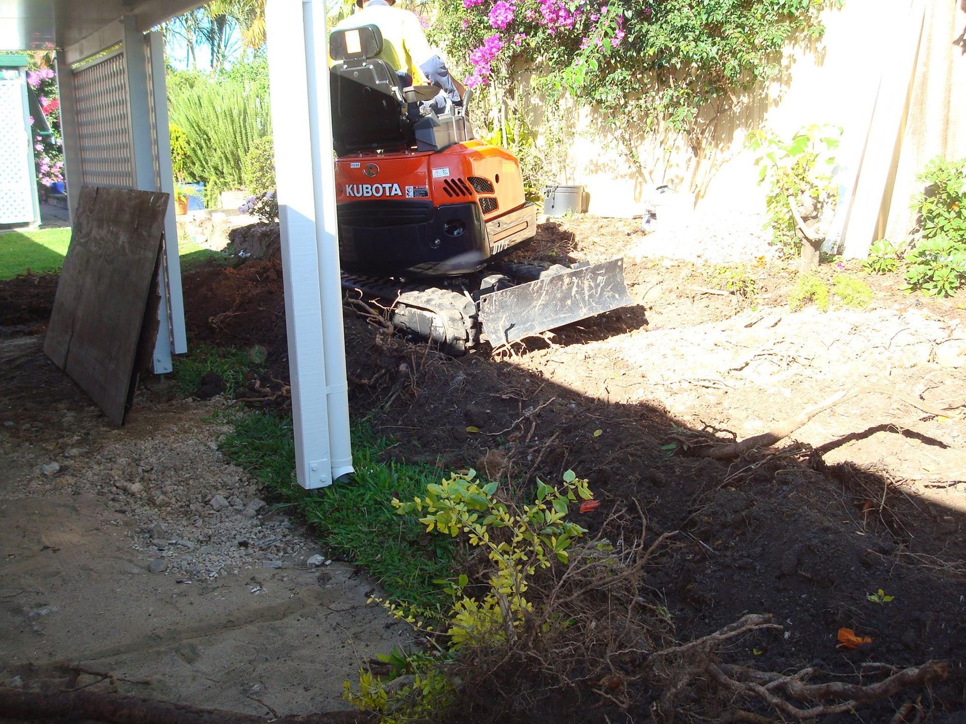 Mini excavator digging in a yard near a covered structure. Dark soil, green plants, and a light-colored structure.