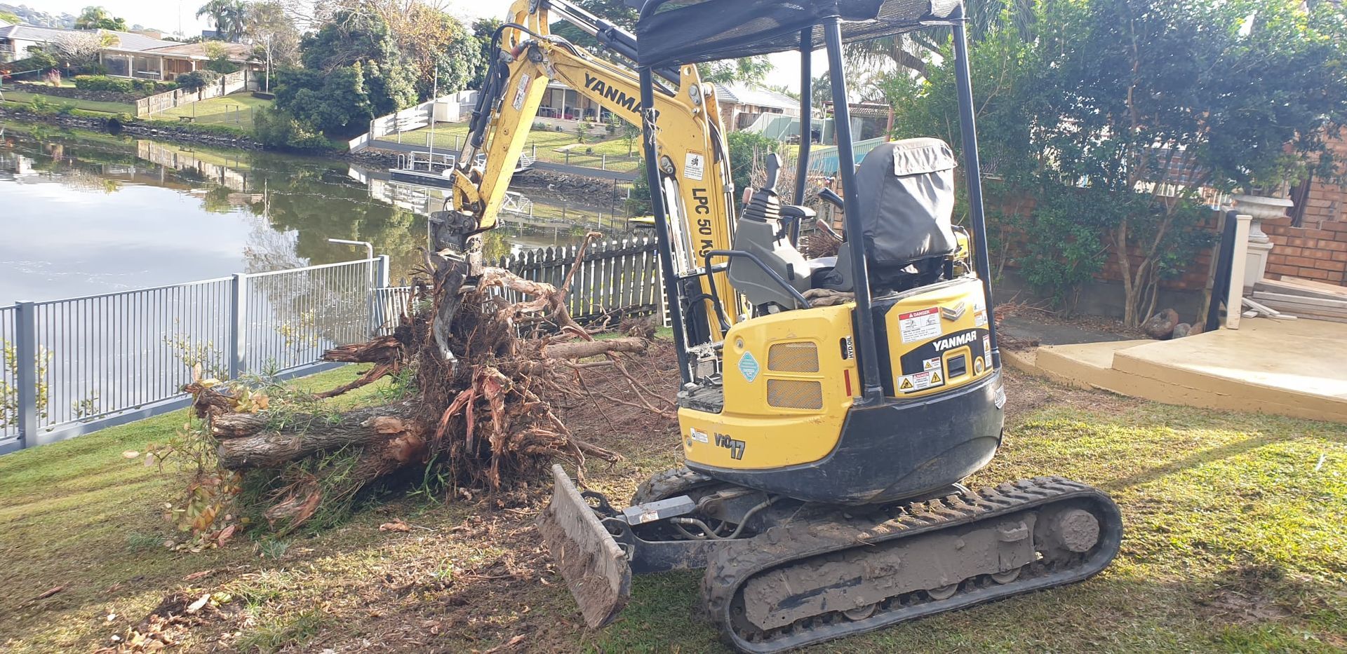 A yellow excavator is removing debris on a grassy bank near a lake.