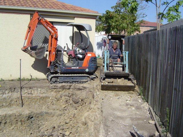 Two small excavators digging a trench next to a fence and house. One has an operator.