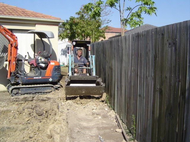 Man operating a small roller machine in a narrow trench next to a wooden fence with an excavator in the background.