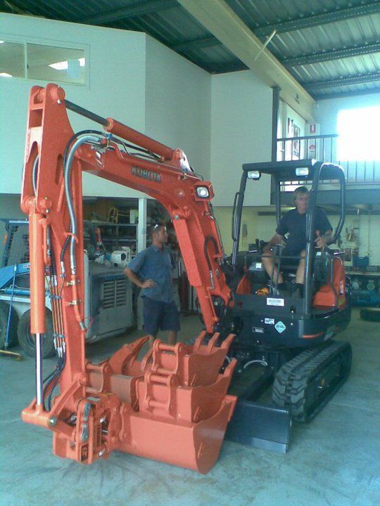 Two men near an orange Kubota excavator indoors. One man sits in the cab, the other stands beside it.