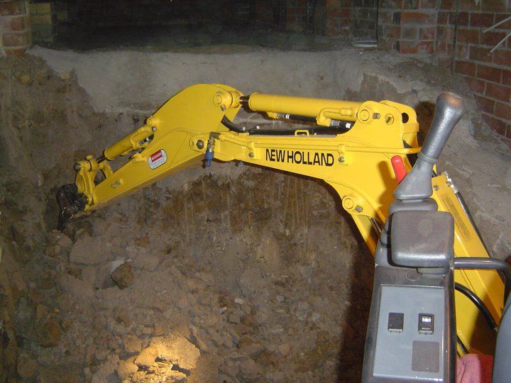 Yellow New Holland mini-excavator digging in an excavation, inside a brick-lined space.