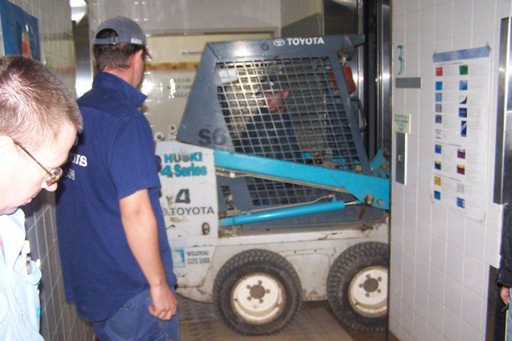A blue Toyota skid steer loader in an elevator, with two men looking on.