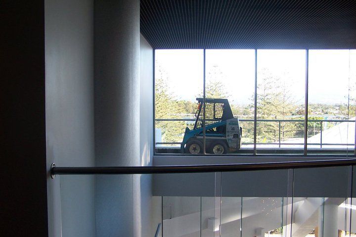 A small, blue forklift sits behind a large window. A building interior with a railing is in the foreground.