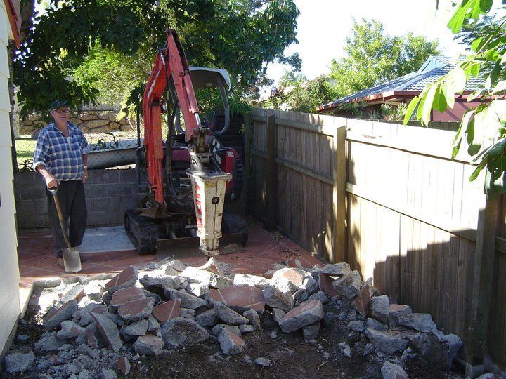 Man using a jackhammer to break up pavement in a backyard; pile of rubble, wooden fence, tree.