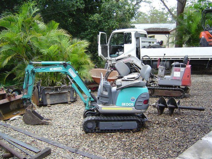 Small teal excavator with bucket and auger next to a smaller red excavator. White truck in the background.