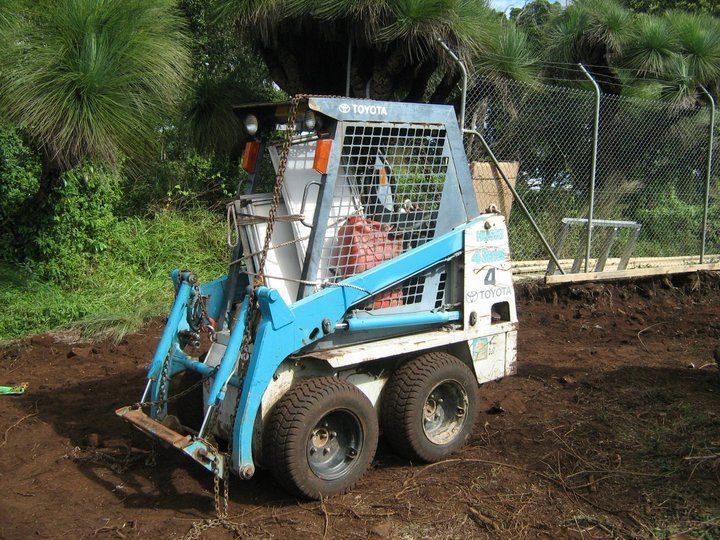 Blue and white skid steer loader on a dirt surface near trees, ready for construction.