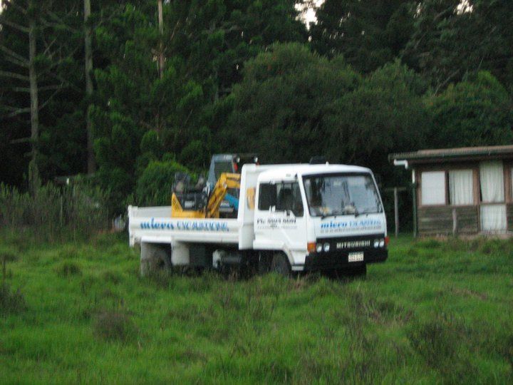 White Mitsubishi truck with mini excavator parked in tall grass near trees and a building.