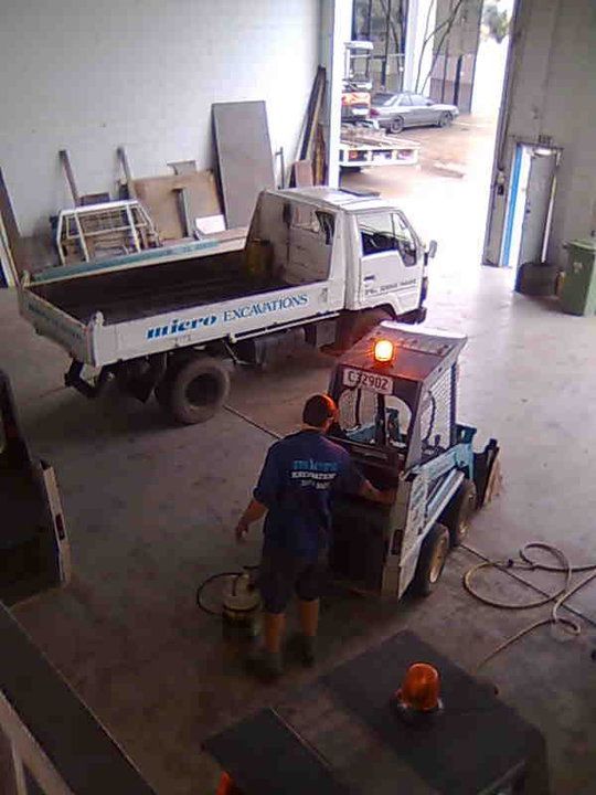 A person operating a small excavator near a white truck in a garage; both have construction logos.