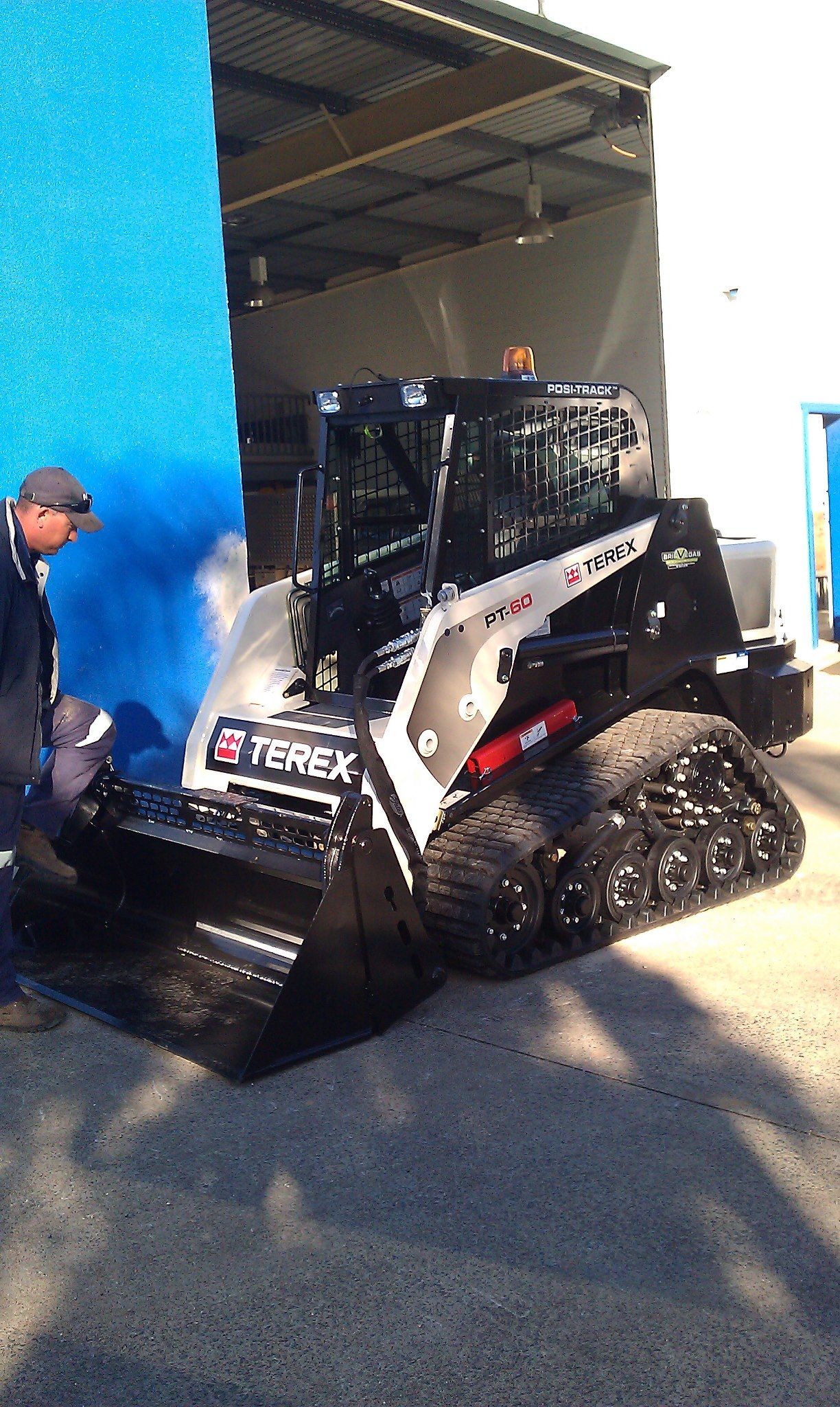 A Terex compact track loader sits on a concrete surface. A person stands nearby, blue and white building in the background.