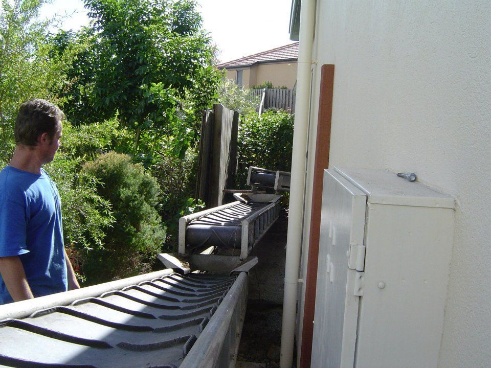 Man looking at a gutter with a makeshift contraption. The contraption is on the side of a building, surrounded by greenery.