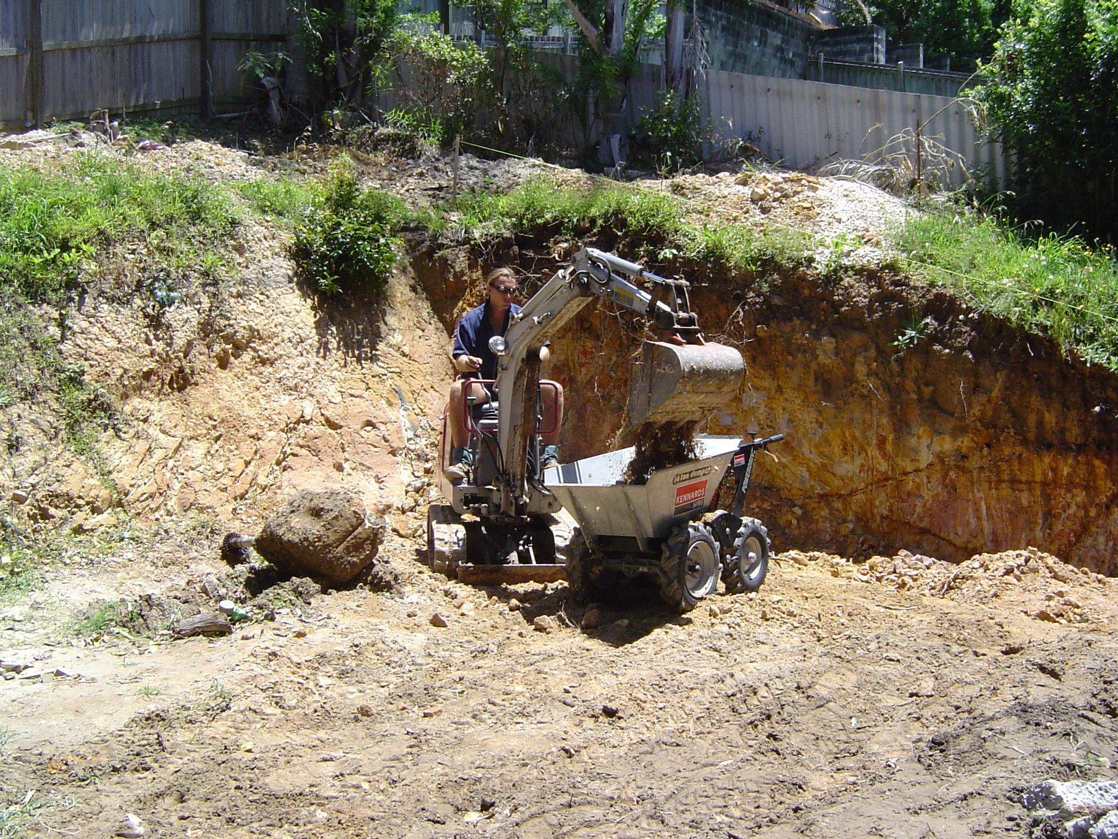 Person operating small excavator, loading dirt into a wheelbarrow in a construction site.