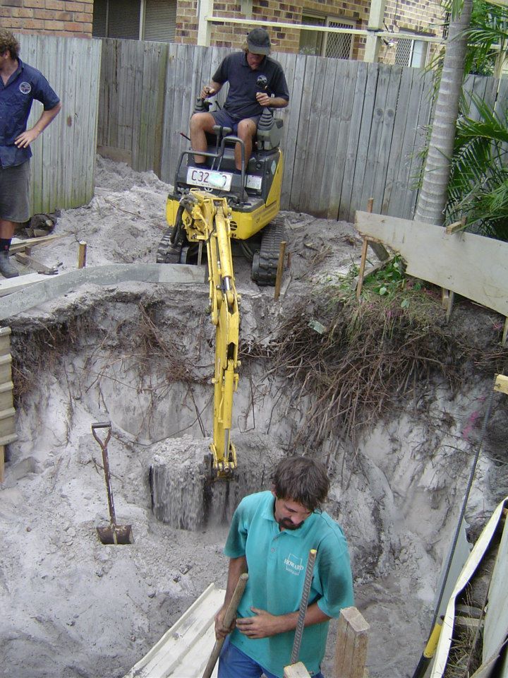 Men operating a small excavator, digging near a fence. One man observes.